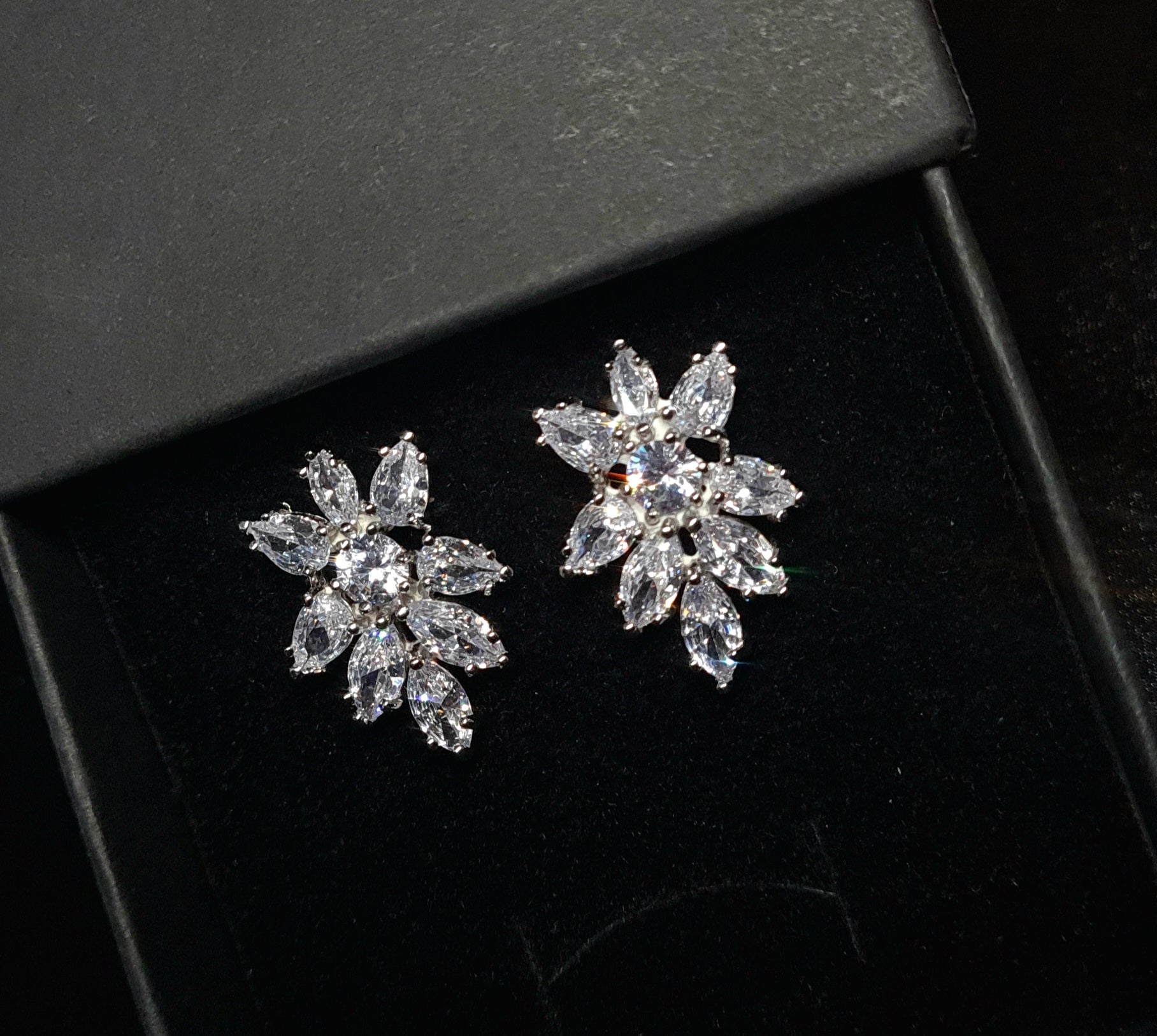 A pair of earrings in the shape of a flower, made from cubic zirconia stones. The earrings are sitting on a black surface. The flower is about 2.6 cm and is made up of petals that are clear and sparkling. The center of the flower is a large cubic zirconia stone.