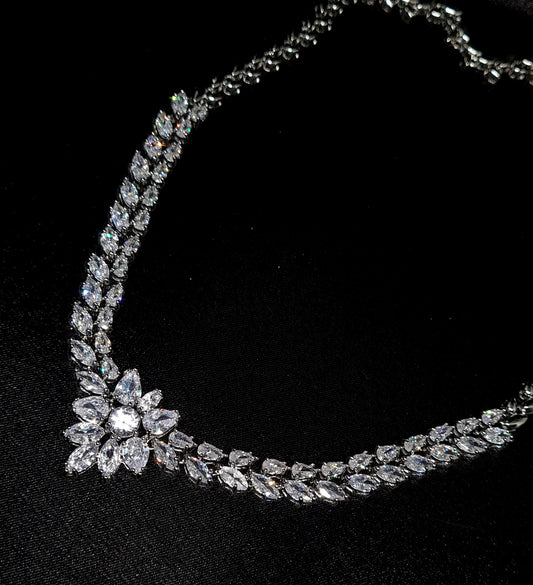 A close-up of a necklace on a black background. The necklace is made of silver and has diamonds in a variety of shapes and sizes. The diamonds are the main focus of the image and they sparkle in the light.