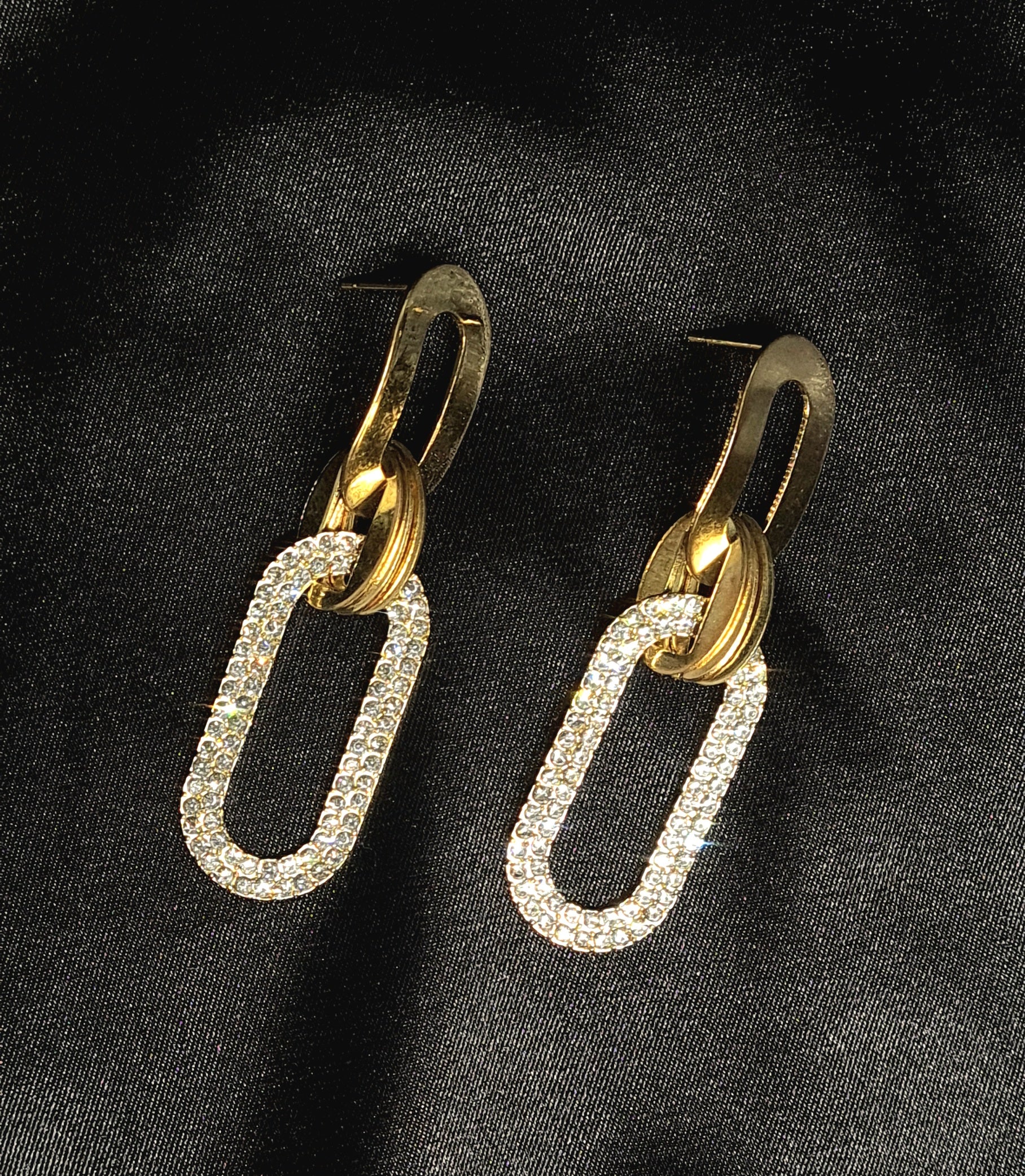 A pair of gold earrings with rhinestones on a black background. The earrings are teardrop-shaped with a gold border. The rhinestones are clear and round. The background is a black velvet cloth.