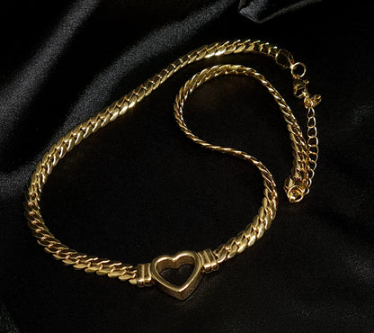 Gold necklace with heart pendant on black background. The necklace is made of gold and has a heart pendant in the center. The heart is gold and has a smooth surface. The necklace is sitting on a black background, which provides a nice contrast to the gold color of the heart