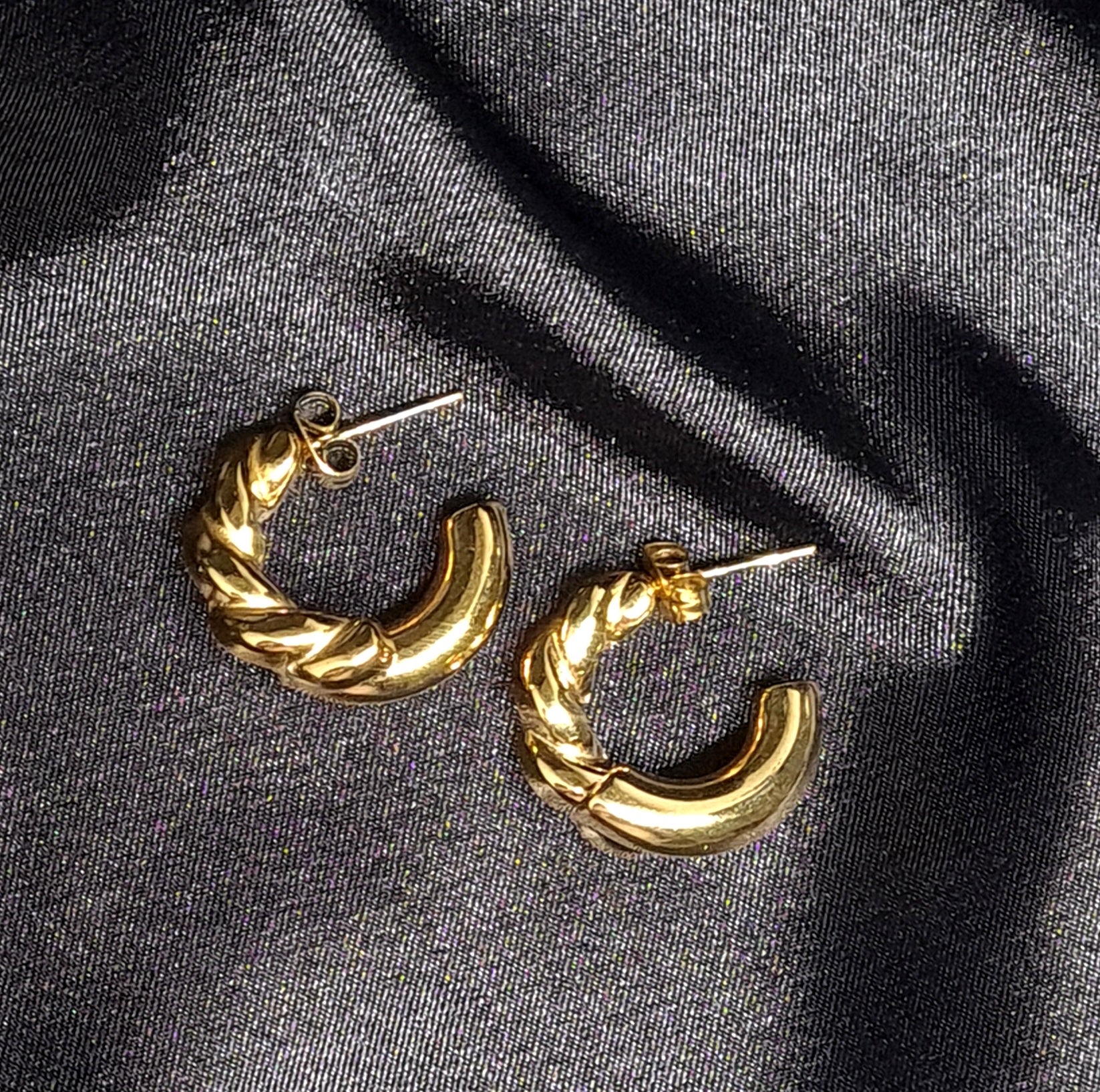 Gold hoop earrings sitting on black cloth. The earrings are a classic and elegant style that would be perfect for any occasion. The earrings are made of gold and have a hoop design. The hoops are large and round, and they sit on a black cloth.
