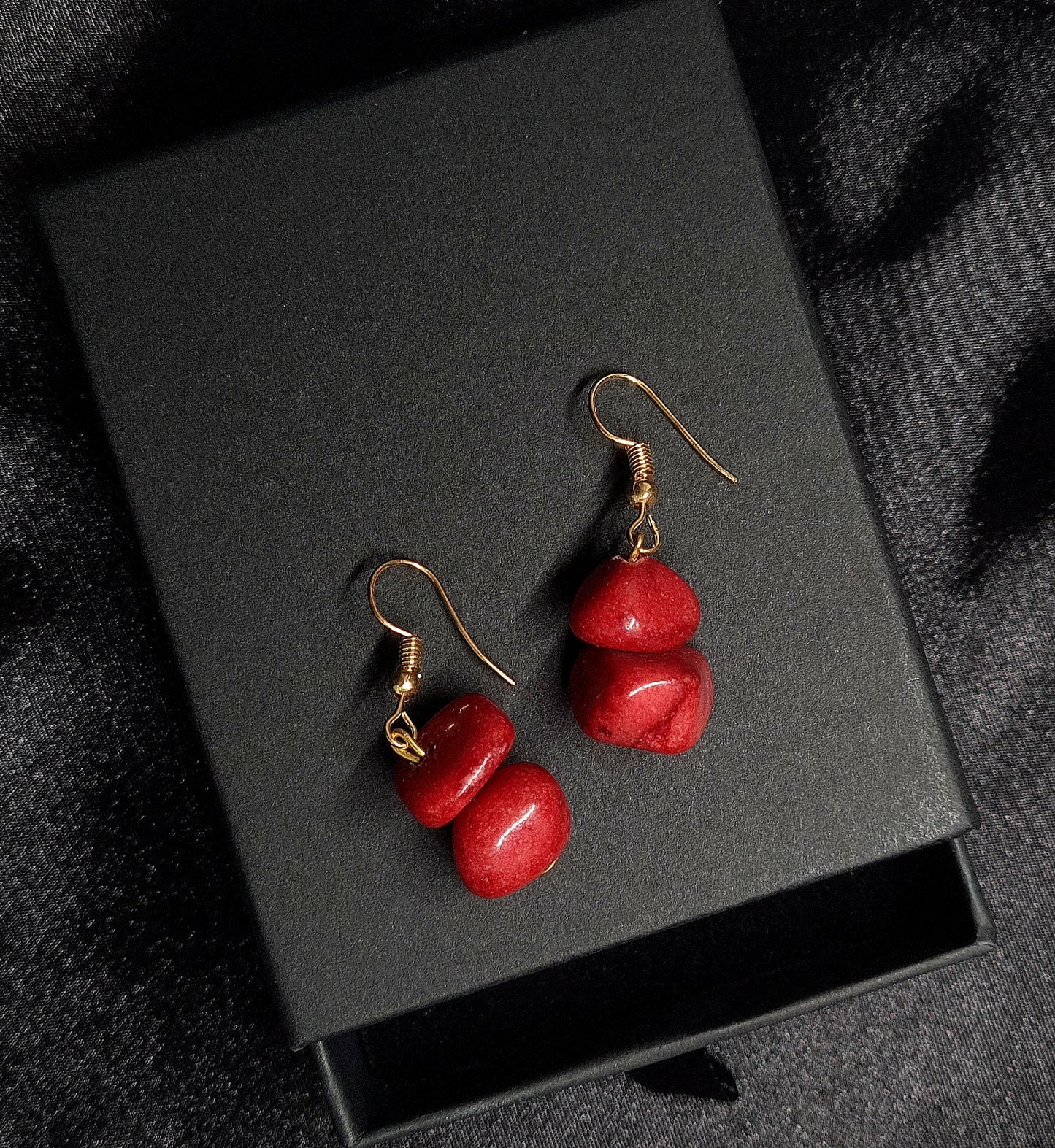 Pair of red earrings sitting on a black box. The earrings are made of red metal and have gold hooks. They are a simple and elegant design that would be perfect for any occasion. The earrings are sitting on a black box, which provides a nice contrast to the red color of the earrings.