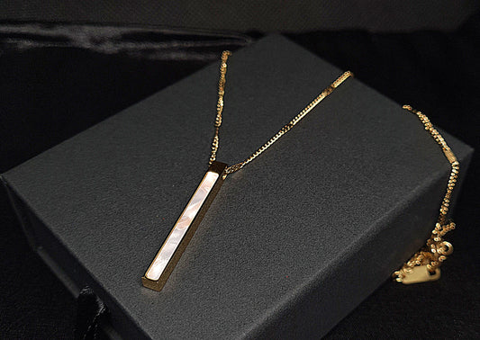 Gold bar necklace on black box. The necklace is made of gold and has a simple bar design. The bar is long and thin, and it has a smooth surface. The necklace is sitting on a black box, which provides a nice contrast to the gold color of the necklace.