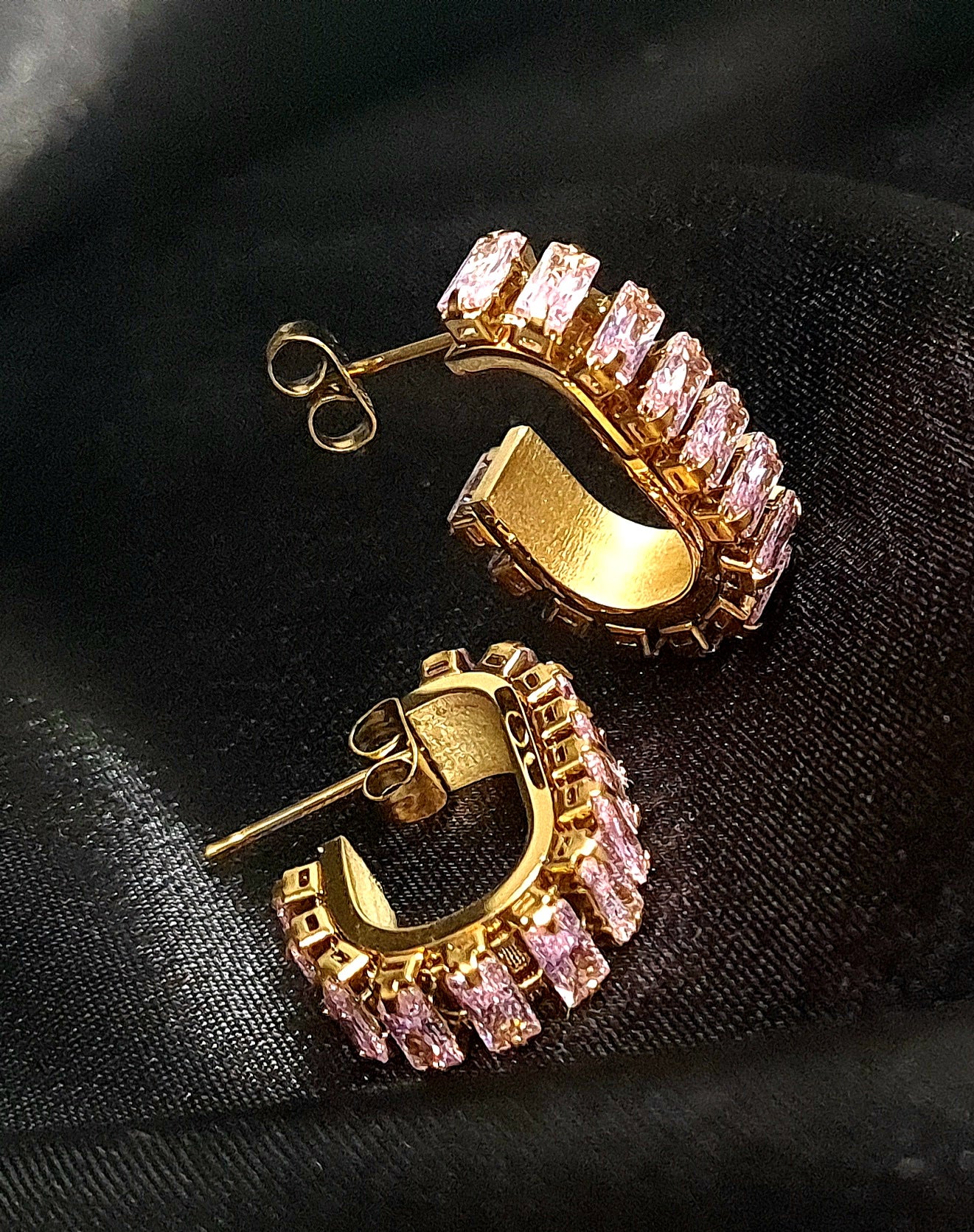 A pair of gold geometric design hoop earrings with pink cubic zirconia stones on a black cloth.