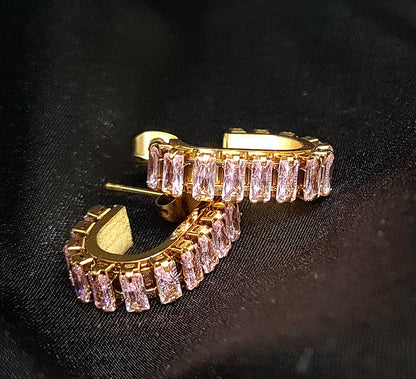 A pair of gold geometric design hoop earrings with pink cubic zirconia stones on a black cloth.
