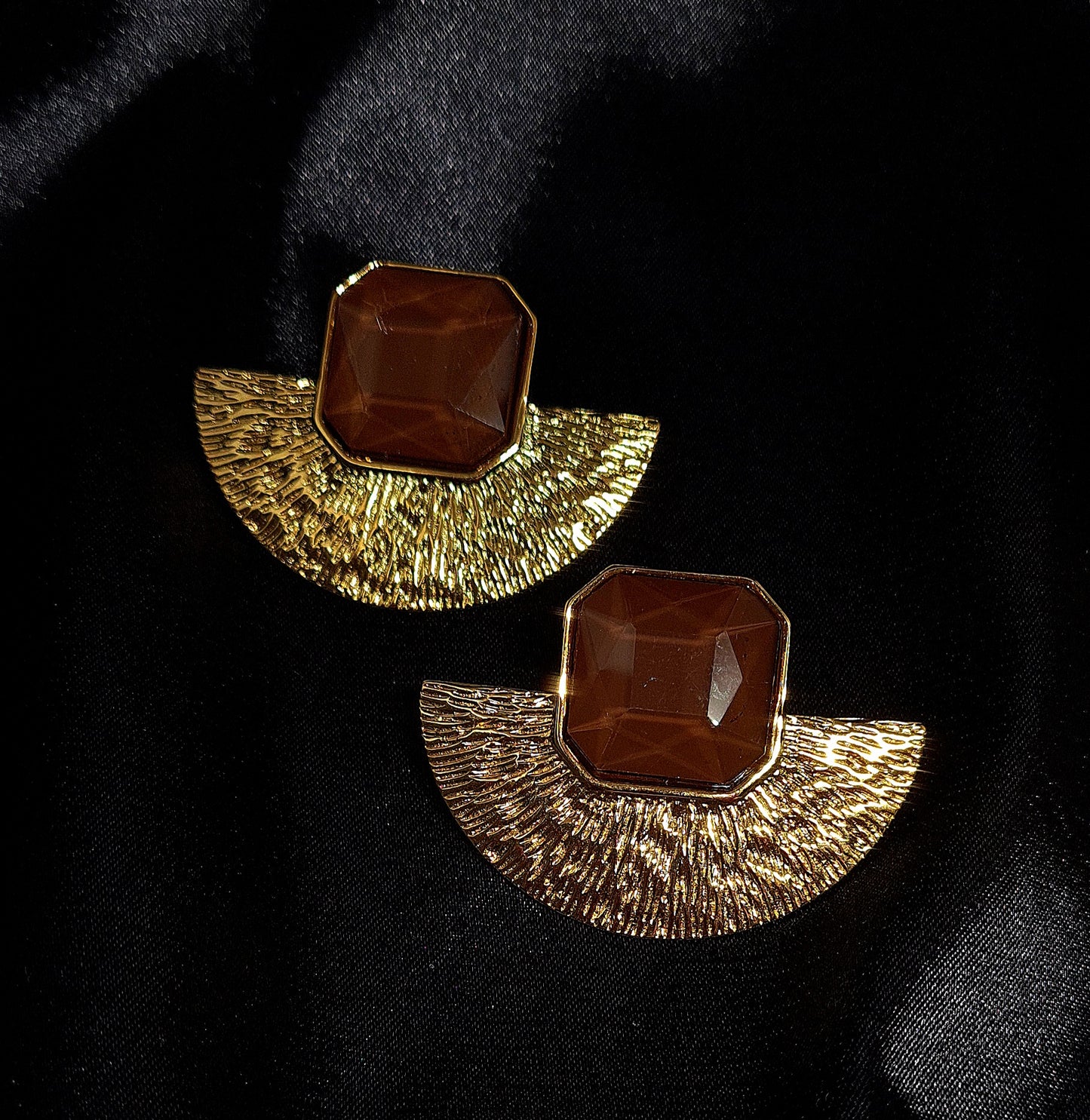A pair of gold earrings with brown stones on a black surface. The earrings are fan-shaped with a delicate design. They are made of gold with brown stones in the center. The earrings are displayed on a black cloth.