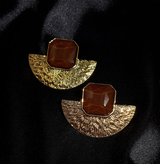 A pair of gold earrings with brown stones on a black surface. The earrings are fan-shaped with a delicate design. They are made of gold with brown stones in the center. The earrings are displayed on a black cloth.