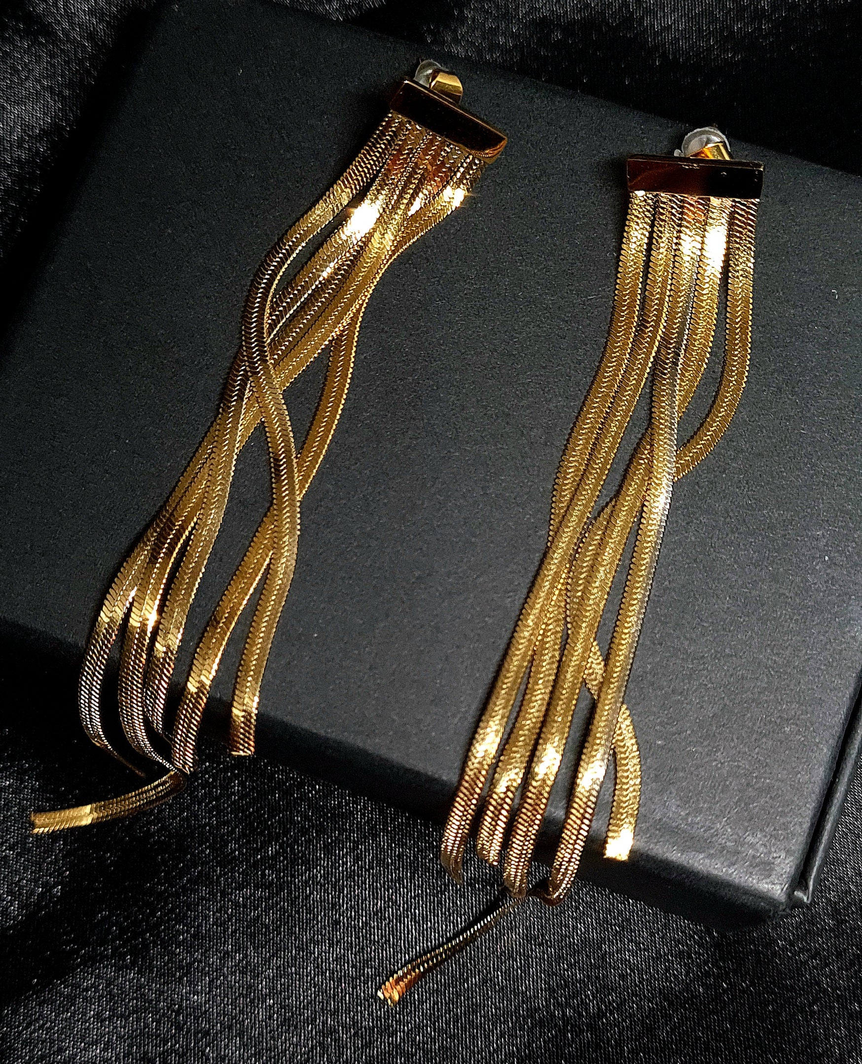 Gold chain earrings sitting on a black box. The earrings are long and have a fringe design. They are made of gold and have a shiny finish. The earrings are sitting on top of a black box.