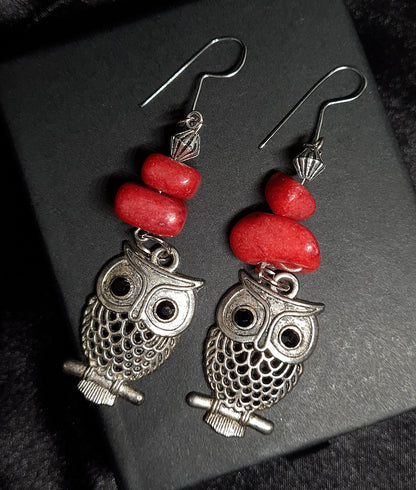 Pair of silver owl earrings with red beads on a black box. The earrings are made of silver and have a realistic owl design. The owl has large eyes, a beak, and wings. The earrings are decorated with red beads. The earrings are sitting on a black box