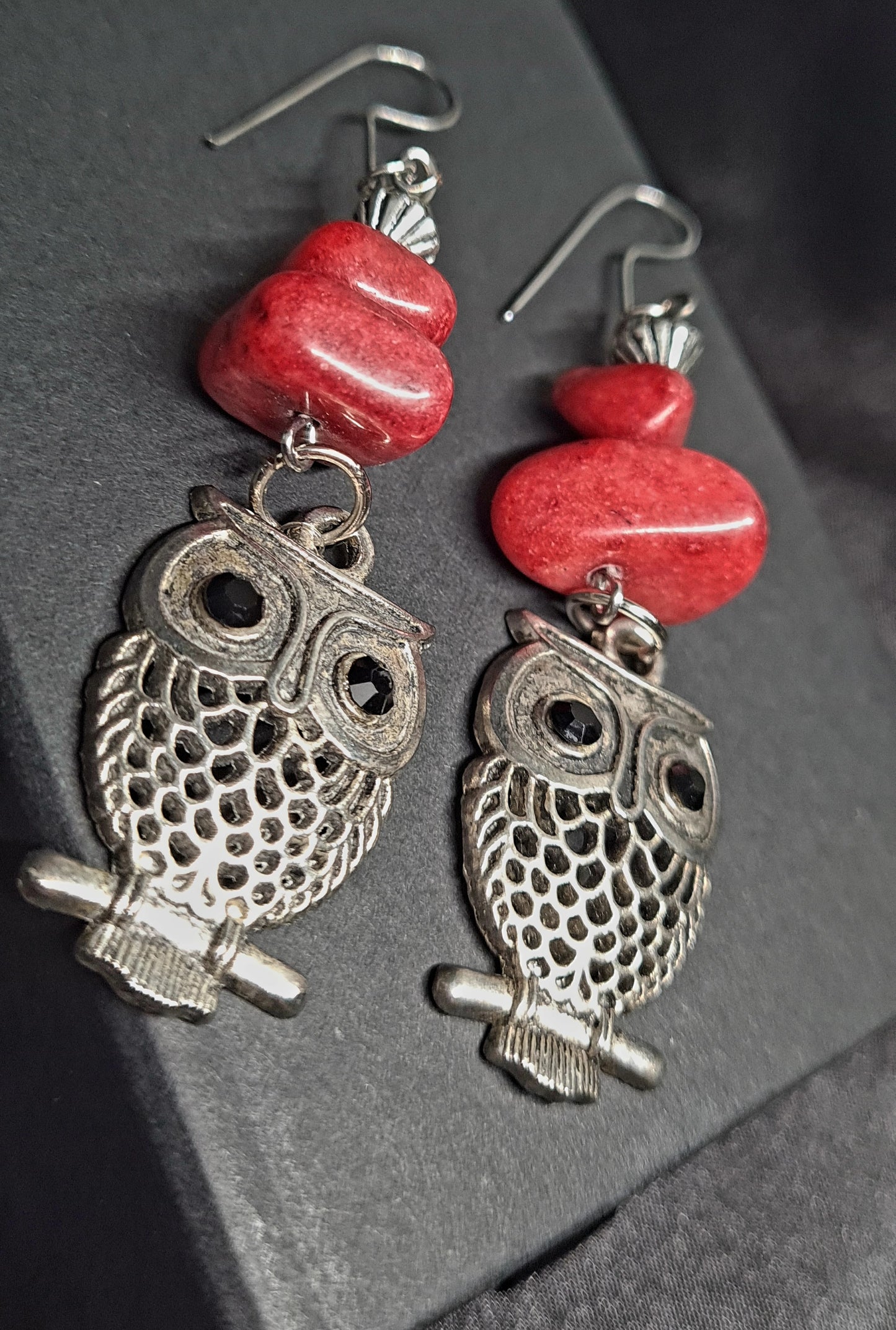 Pair of silver owl earrings with red beads on a black box. The earrings are made of silver and have a realistic owl design. The owl has large eyes, a beak, and wings. The earrings are decorated with red beads. The earrings are sitting on a black box
