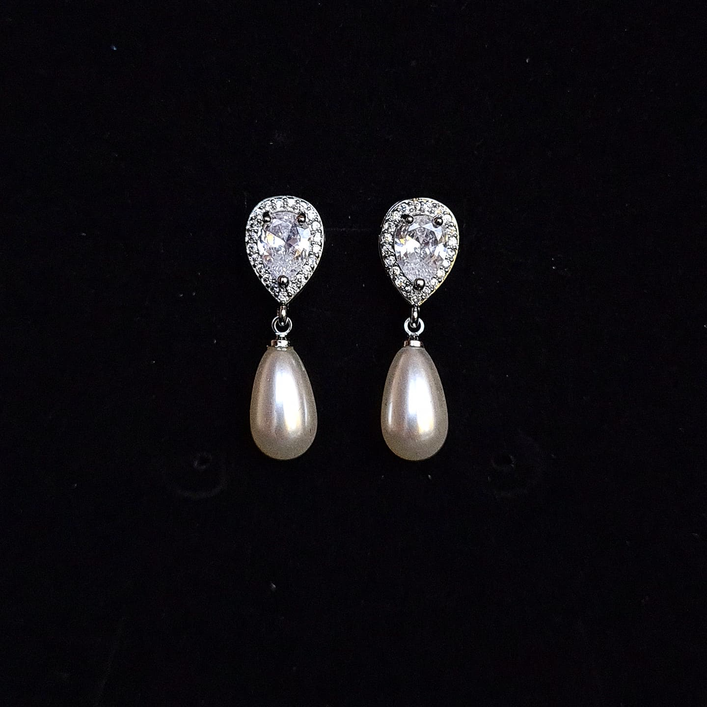 A pair of silver earrings with pearls and diamonds on a black box. The earrings are teardrop-shaped and have a simple, elegant design. The pearls are white and the diamonds are clear. 