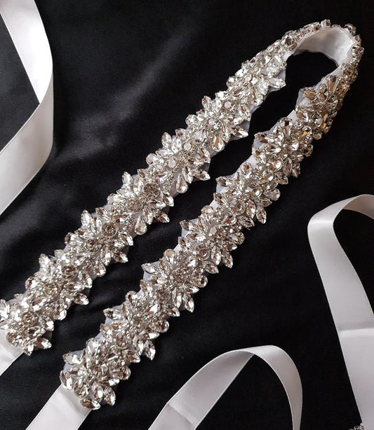 A bridal belt with rhinestones and a white ribbon on a black background. The belt is made of satin and has a intricate design with rows of rhinestones. The rhinestones are sparkling and the belt is very shiny. The white ribbon adds a touch of elegance.