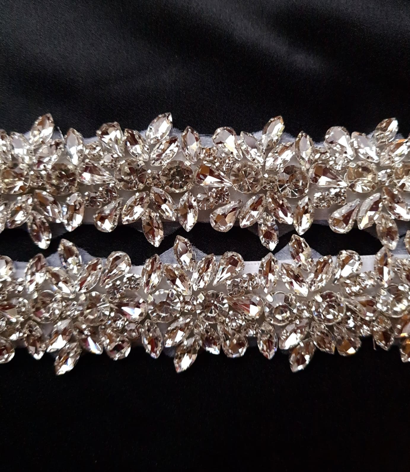 a close up view of A bridal belt with rhinestones and a white ribbon on a black background. The belt is made of satin and has a intricate design with rows of rhinestones. The rhinestones are sparkling and the belt is very shiny. The white ribbon adds a touch of elegance.