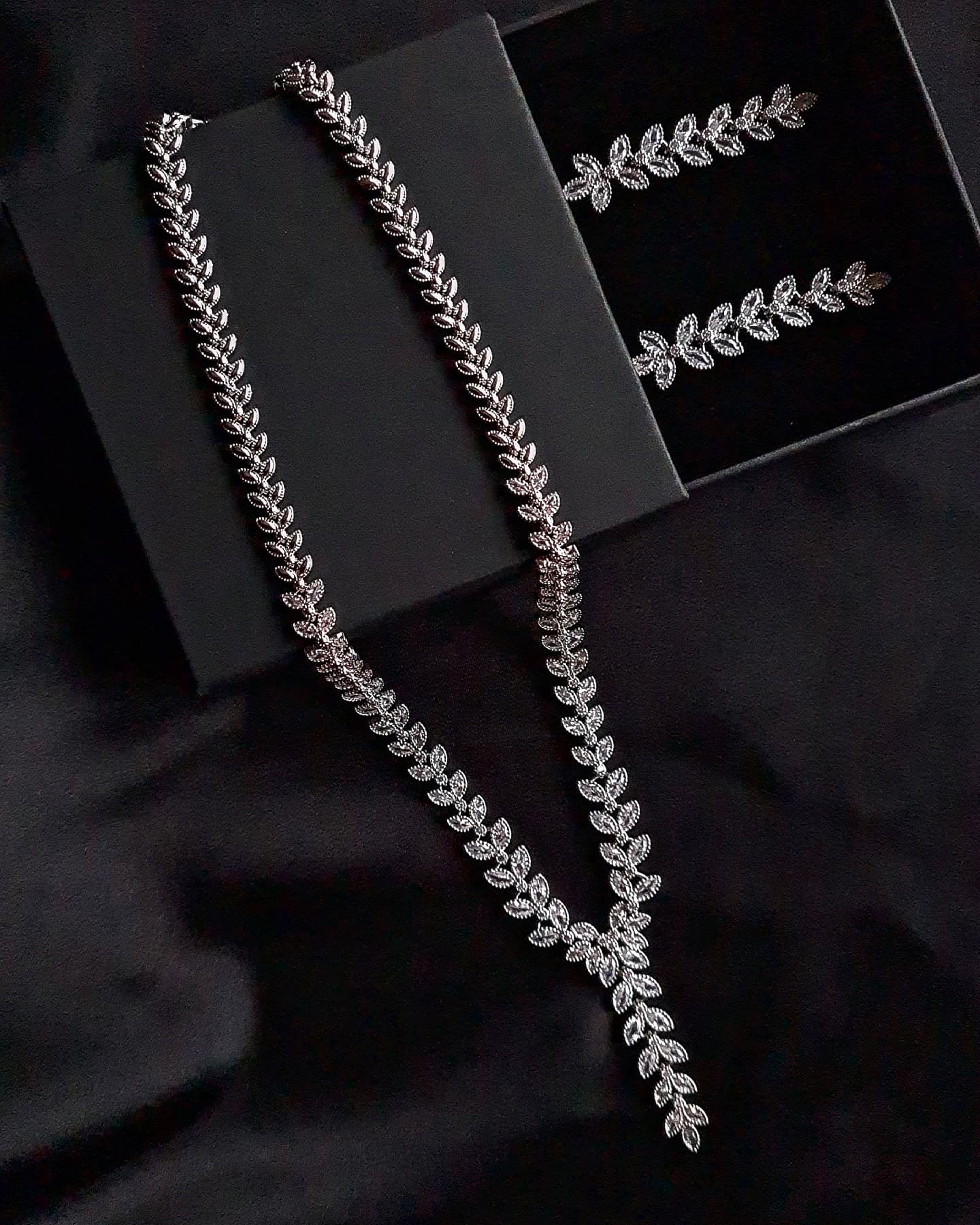 Overhead view of a silver necklace sparkling with floral shaped Cubic Zirconia diamonds in a variety of shapes and sizes. and a pair of a floral shape mini earrings studded with white crystals displayed in a black jewelry box.