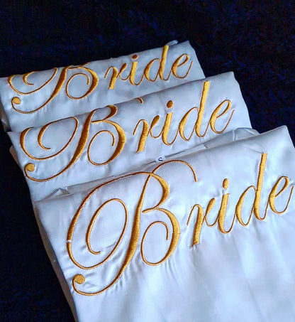 overhead shot of 3 white kimonos with the word "bride" embroidered on it. The kimono is made of silk and has a simple design. The word "bride" is embroidered in gold thread and is located in the center of the kimono. The kimono is very elegant and would be perfect for a wedding.