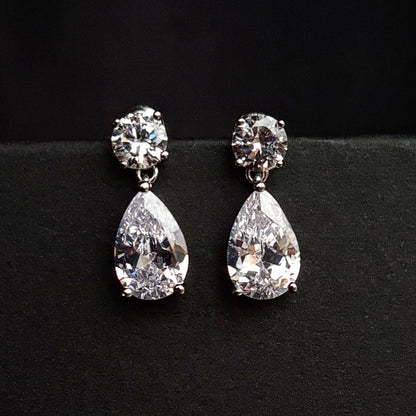 A pair of silver earrings with diamond studs on a black surface. The earrings are teardrop-shaped and have a simple, elegant design. The diamonds are clear and sparkling.