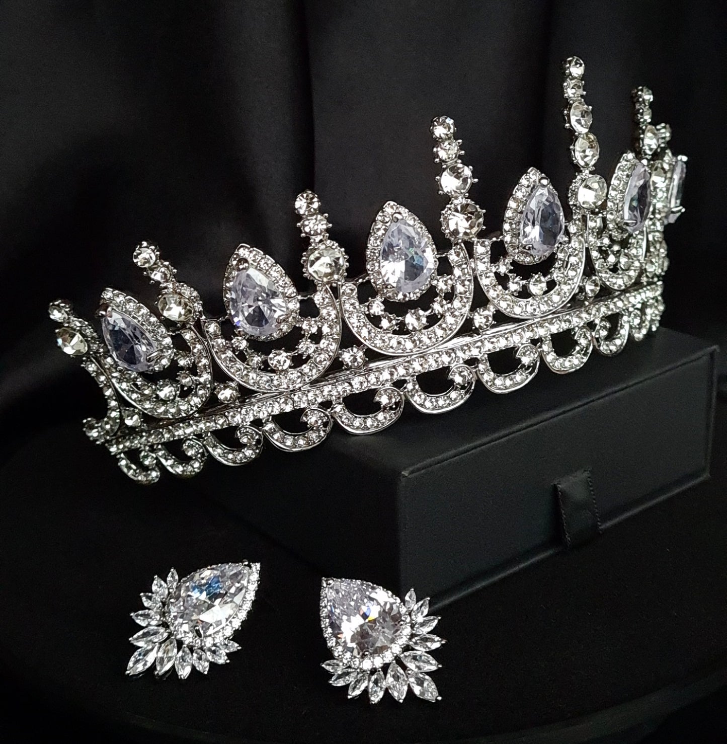 A tiara and earrings set sitting on a black box. The tiara is made of silver and has a delicate design with crystals and cubic zirconia. The earrings are also made of silver and have a matching design.