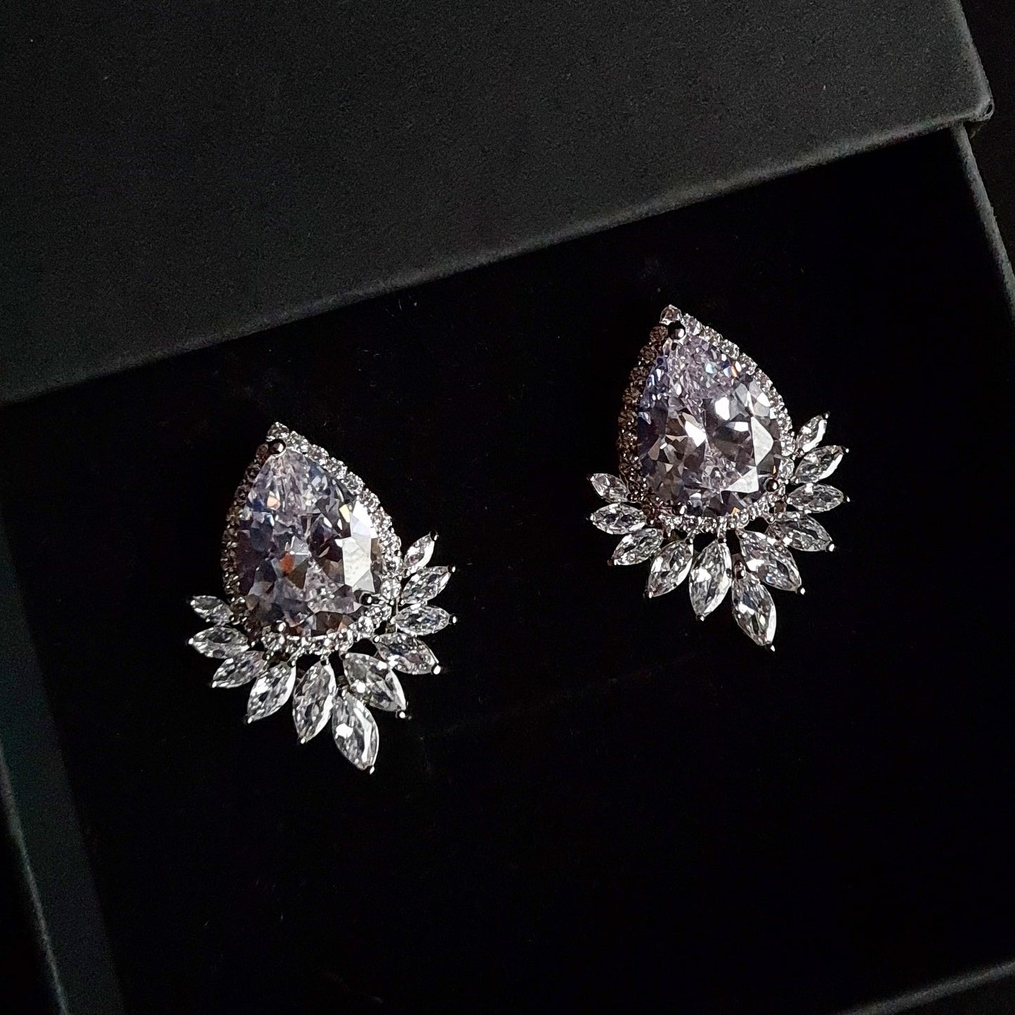 A pair of silver earrings with a large diamond in the center and a cluster of smaller diamonds around it. The earrings are sitting inside a black jewelry box.