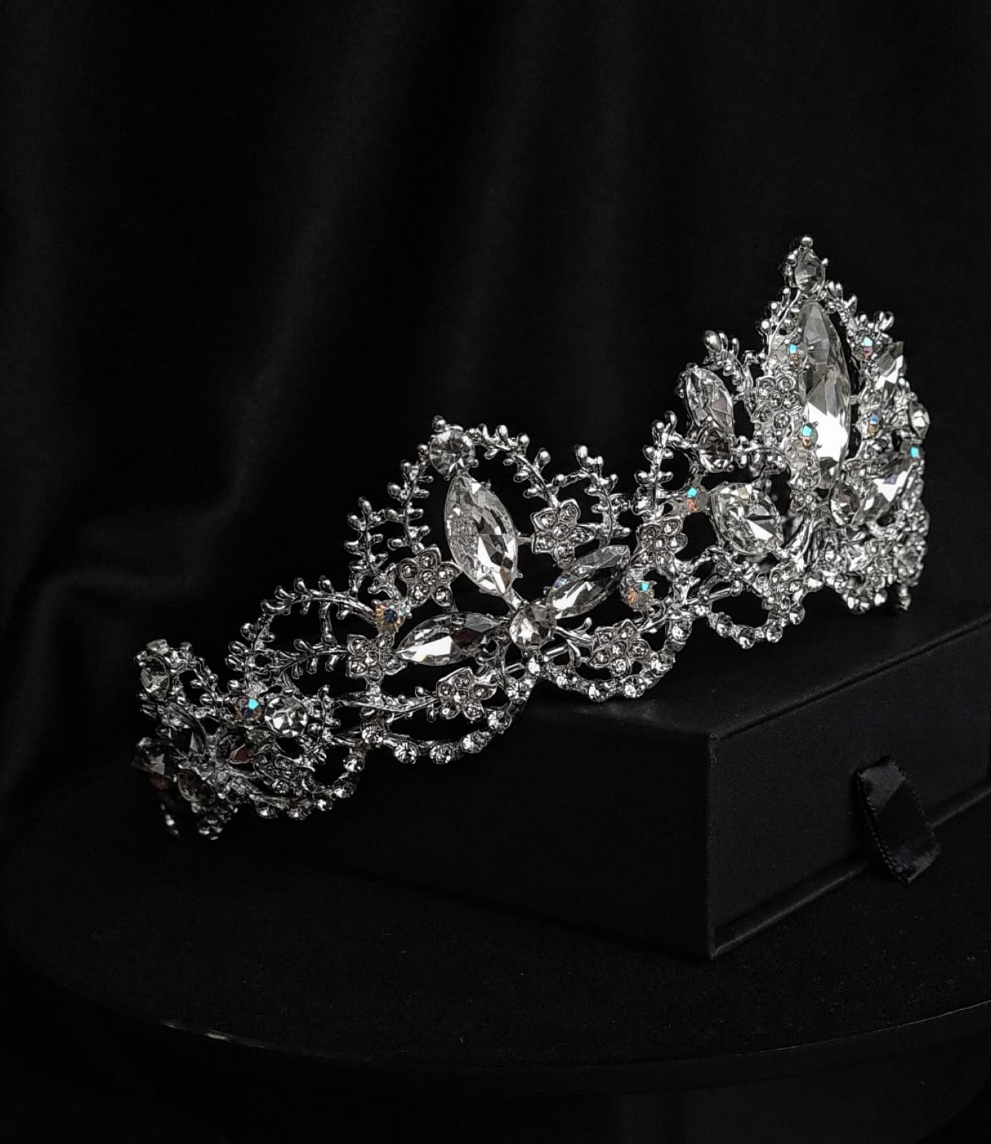  A tiara with rhinestones on a black box. The tiara is made of silver and features a delicate design with cascading rhinestones. The rhinestones are clear and sparkling. The tiara is sitting on top of a black box.