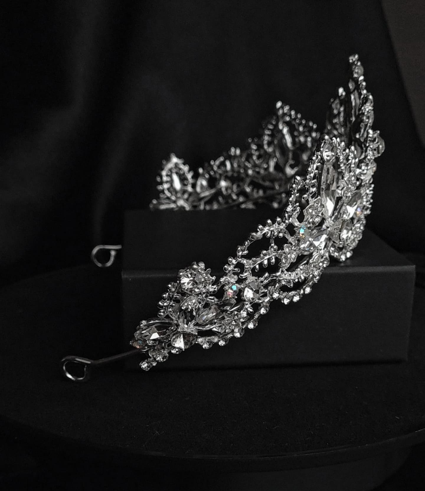 side view of  A tiara with rhinestones on a black box. The tiara is made of silver and features a delicate design with cascading rhinestones. The rhinestones are clear and sparkling. The tiara is sitting on top of a black box.
