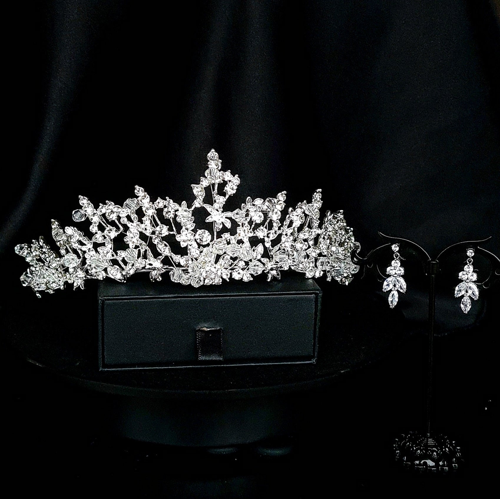 A silver tiara with rhinestones sitting on top of a black box. The tiara is made up of multiple curved bands, each of which is decorated with rhinestones. The tiara is sitting on top of a black box, which is partially obscuring the bottom of the tiara. next to an earrings
