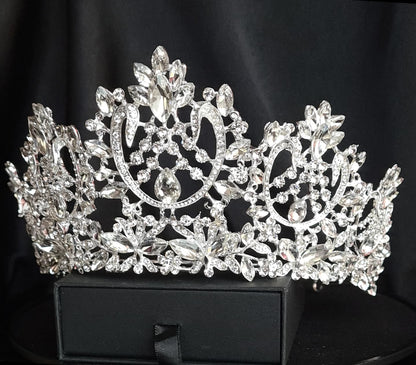 A silver tiara with a teardrop-shaped center stone and a design of intertwined vines and flowers. The tiara is encrusted with clear cubic zirconia stones. The tiara is sitting on a black velvet surface.
