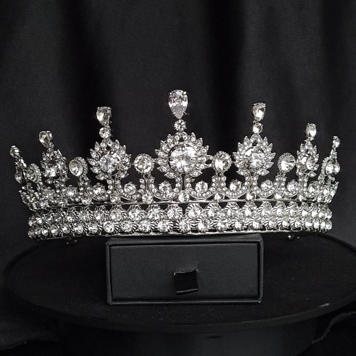 A close-up of a tiara with rhinestones on a black background. The tiara is made of silver and features a delicate design with cascading rhinestones. The rhinestones are clear and sparkling.