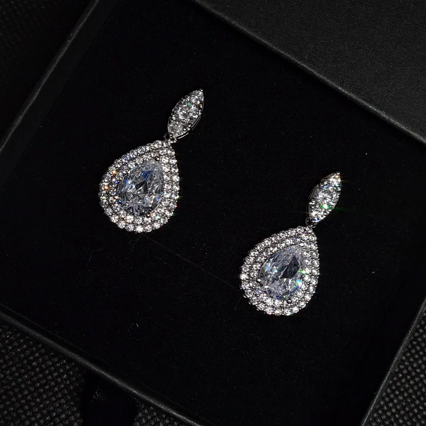 A pair of diamond earrings on a black background. The earrings are made of silver and are set with round cubic zirconia stones. The diamonds are arranged in a cluster and are surrounded by smaller diamonds. The earrings are suspended from a black ear wire. The earrings are elegant and sophisticated, and they would be a perfect addition to any formal outfit.