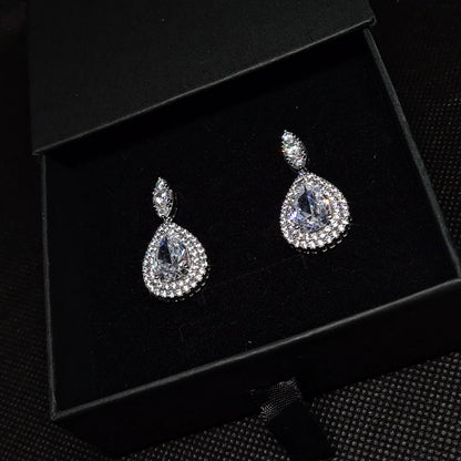 A pair of diamond earrings on a black background. The earrings are made of silver and are set with round cubic zirconia stones. The diamonds are arranged in a cluster and are surrounded by smaller diamonds. The earrings are suspended from a black ear wire. The earrings are elegant and sophisticated, and they would be a perfect addition to any formal outfit.