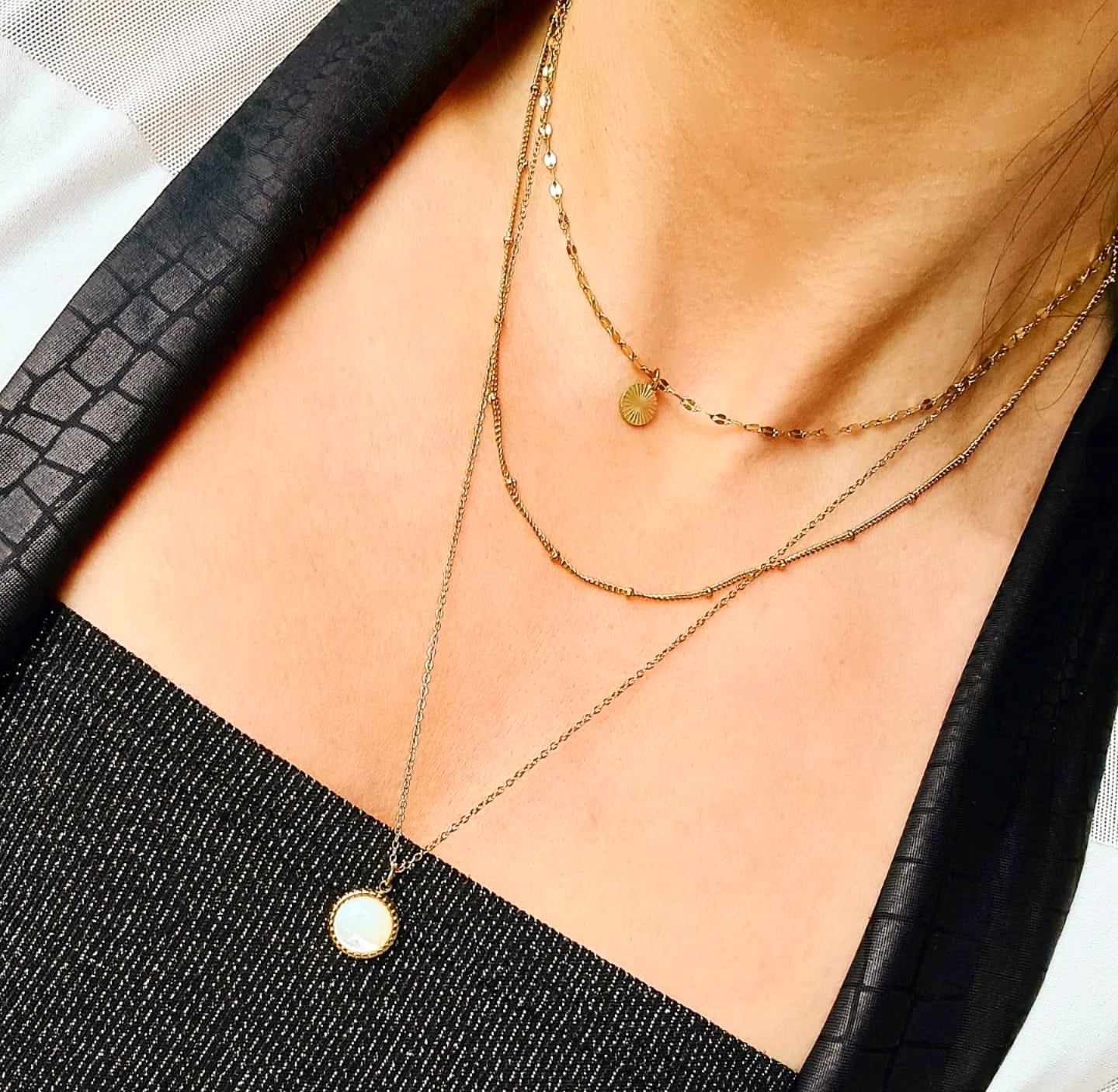 Woman wearing multiple necklaces. The woman is wearing a black top and a gold necklace with a white pendant. She also has a necklace with a stone, and a necklace with a round pendant, and. The necklaces are layered around her neck and add a touch of elegance to her outfit.