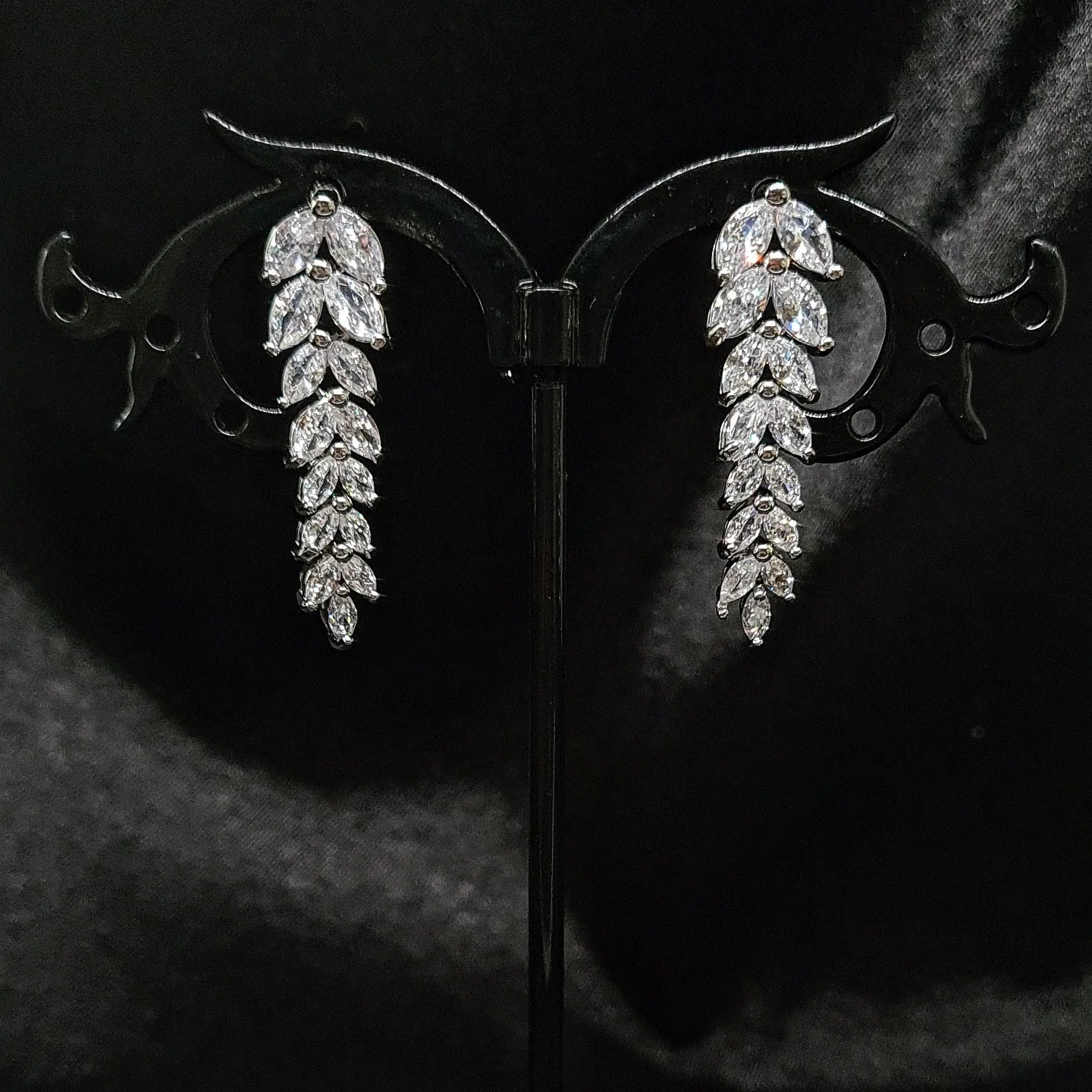 A pair of dangle earrings made of cubic zirconia. The earrings are teardrop-shaped and have a simple, elegant design. The cubic zirconia stones are clear and sparkling. The earrings are sitting on a black cloth.