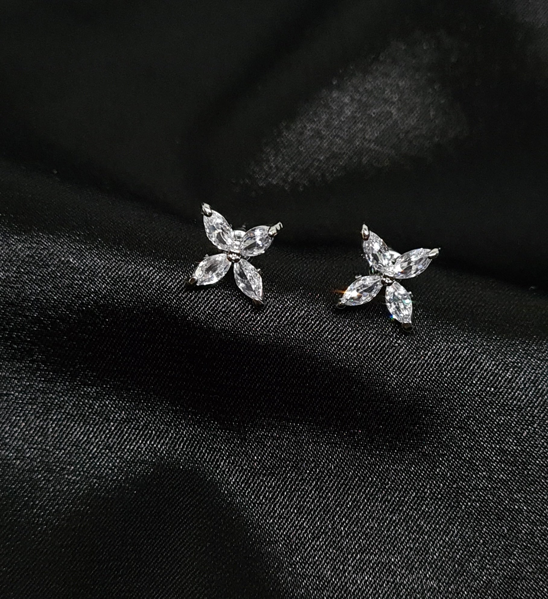 A pair of stud earrings in the shape of a delicate flower, made of cubic zirconia. The earrings are sitting on a black surface. The flower is about 1.2 cm in diameter and is made up of petals that are clear and sparkling. The center of the flower is a small cubic zirconia stone.
