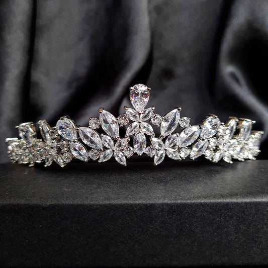 A tiara with diamonds on a black background. The tiara is made of silver and features a delicate design with cascading diamonds. The diamonds are clear and sparkling. The tiara is sitting on a table.