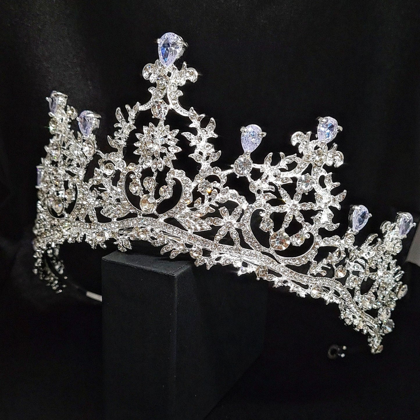  A silver tiara with rhinestones and flowers sitting on a black box. The tiara is made up of multiple curved bands, each of which is decorated with rhinestones and flowers. The tiara is sitting on top of a black box, which is partially obscuring the bottom of the tiara.