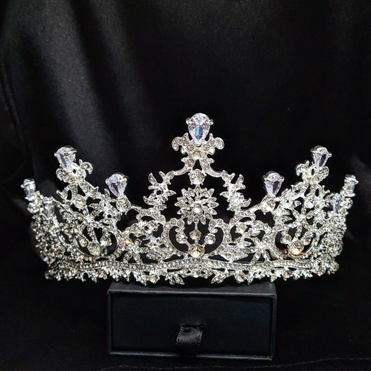  A silver tiara with rhinestones and flowers sitting on a black box. The tiara is made up of multiple curved bands, each of which is decorated with rhinestones and flowers. The tiara is sitting on top of a black box, which is partially obscuring the bottom of the tiara.