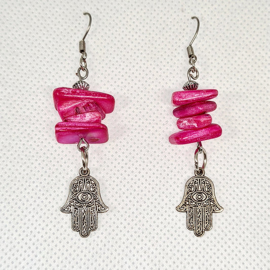 Pair of pink earrings with hamsa pendant. The earrings are made of pink metal and have a hamsa pendant in the center. The hamsa is a hand-shaped amulet that is believed to bring good luck and protection. The earrings are hanging from gold hooks.