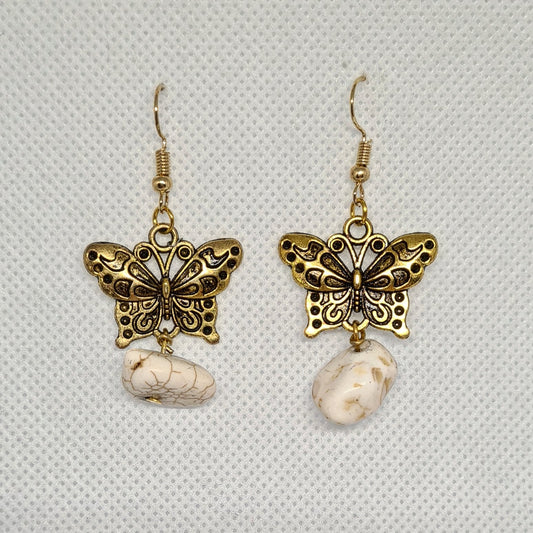 Pair of gold butterfly earrings with white beads on a white background. The earrings are made of gold and have a butterfly design. The butterflies are small and delicate, with white beads for wings. The earrings are hanging from gold hooks. The earrings are sitting on a white background, which allows the details of the earrings to be seen clearly.