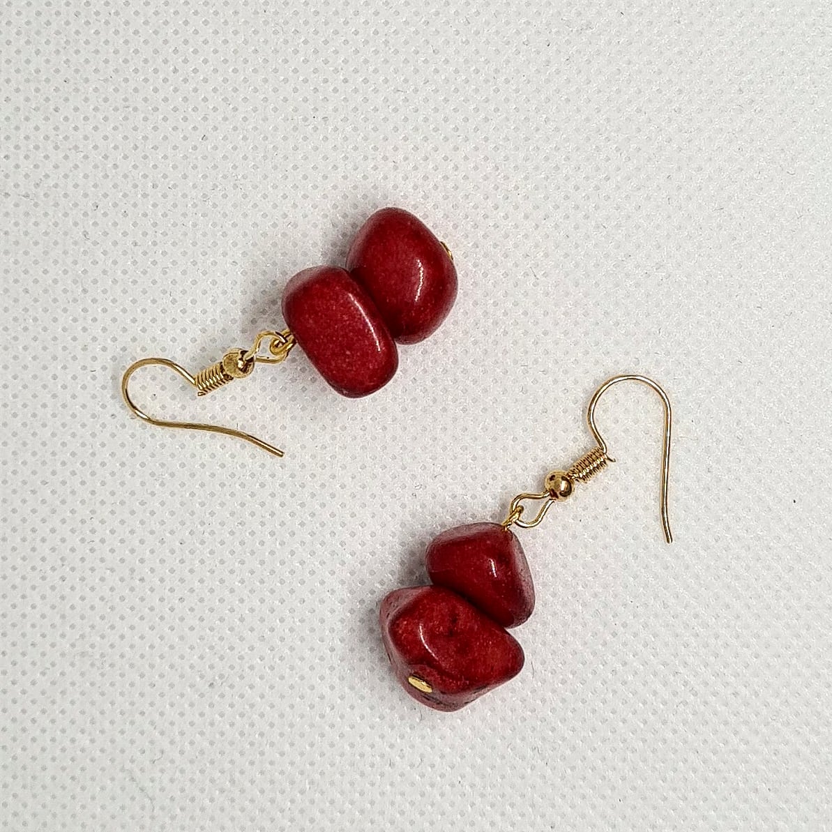 Pair of red earrings sitting on a white surface. The earrings are made of red metal and have gold hooks. They are a simple and elegant design that would be perfect for any occasion. The earrings are sitting on a white surface, which provides a nice contrast to the red color of the earrings.