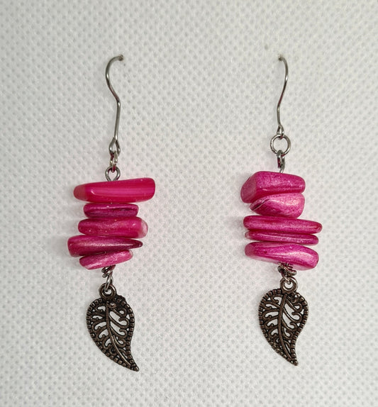 Pair of pink earrings with leaf pendant on a white surface. The earrings are made of pink beads and have a leaf pendant in the center. The leaf is green and has a serrated edge. The earrings are hanging from silver hooks. The earrings are sitting on a white surface.