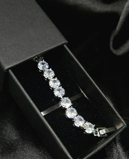 A close-up of a silver bracelet with a single row of diamonds on a black background. The bracelet is simple in design and the diamonds are sparkling.